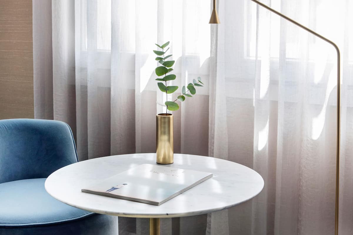 Una habitación moderna con una mesa redonda blanca, dos sillones azules, un jarrón dorado con una planta y una lámpara de pie.