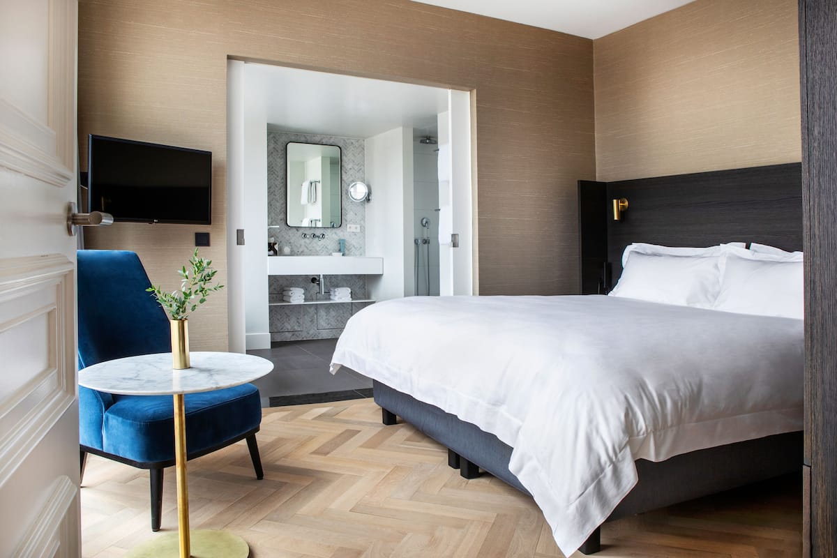 Una habitación de hotel moderna con una cama grande, un televisor montado en la pared, una silla azul y una mesita con una planta.