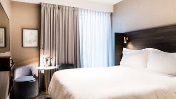 Una habitación de hotel con una cama grande, una mesa redonda con un florero, una silla y un ventanal con cortinas.
