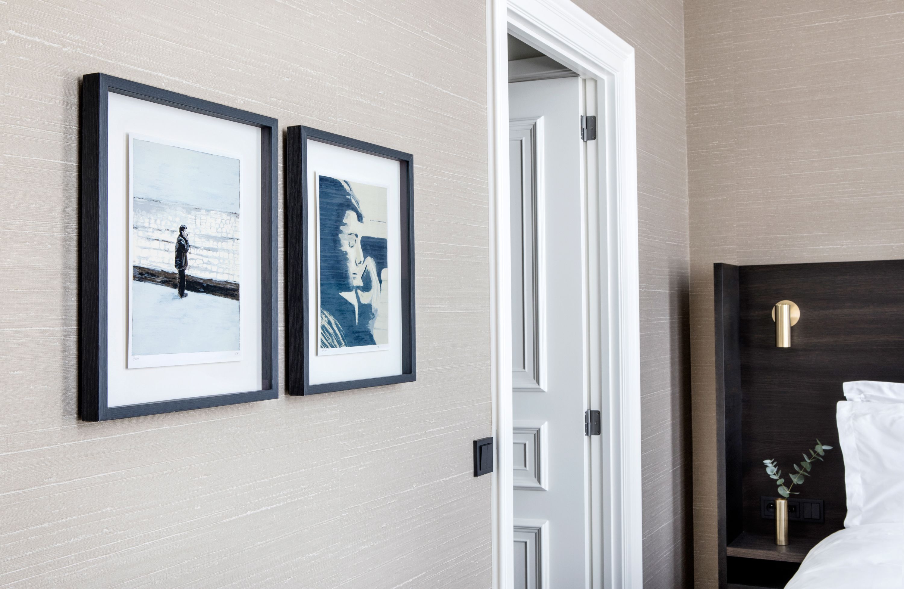 A hotel room with a bed, a bedside table with a lamp, and two framed artworks on the wall.