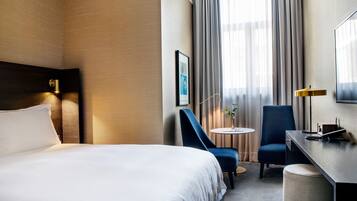 Quarto de hotel com uma cama grande, uma escrivaninha, uma cadeira, uma mesa pequena e um abajur.