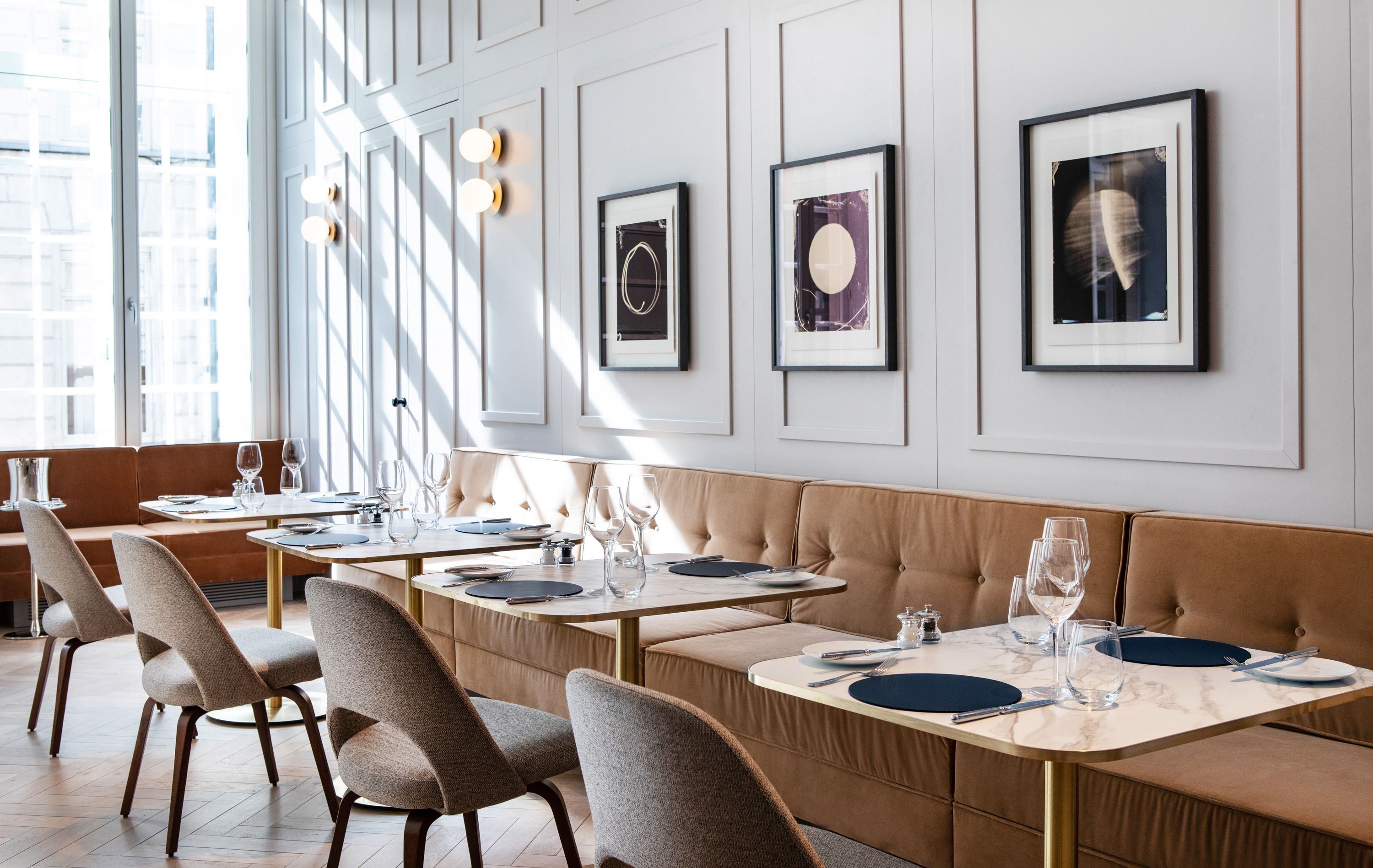 Un'elegante sala da pranzo con una lunga panca, tavoli apparecchiati con piatti, bicchieri e posate. Alle pareti si trovano opere d'arte incorniciate e un'ampia finestra che lascia entrare luce naturale.