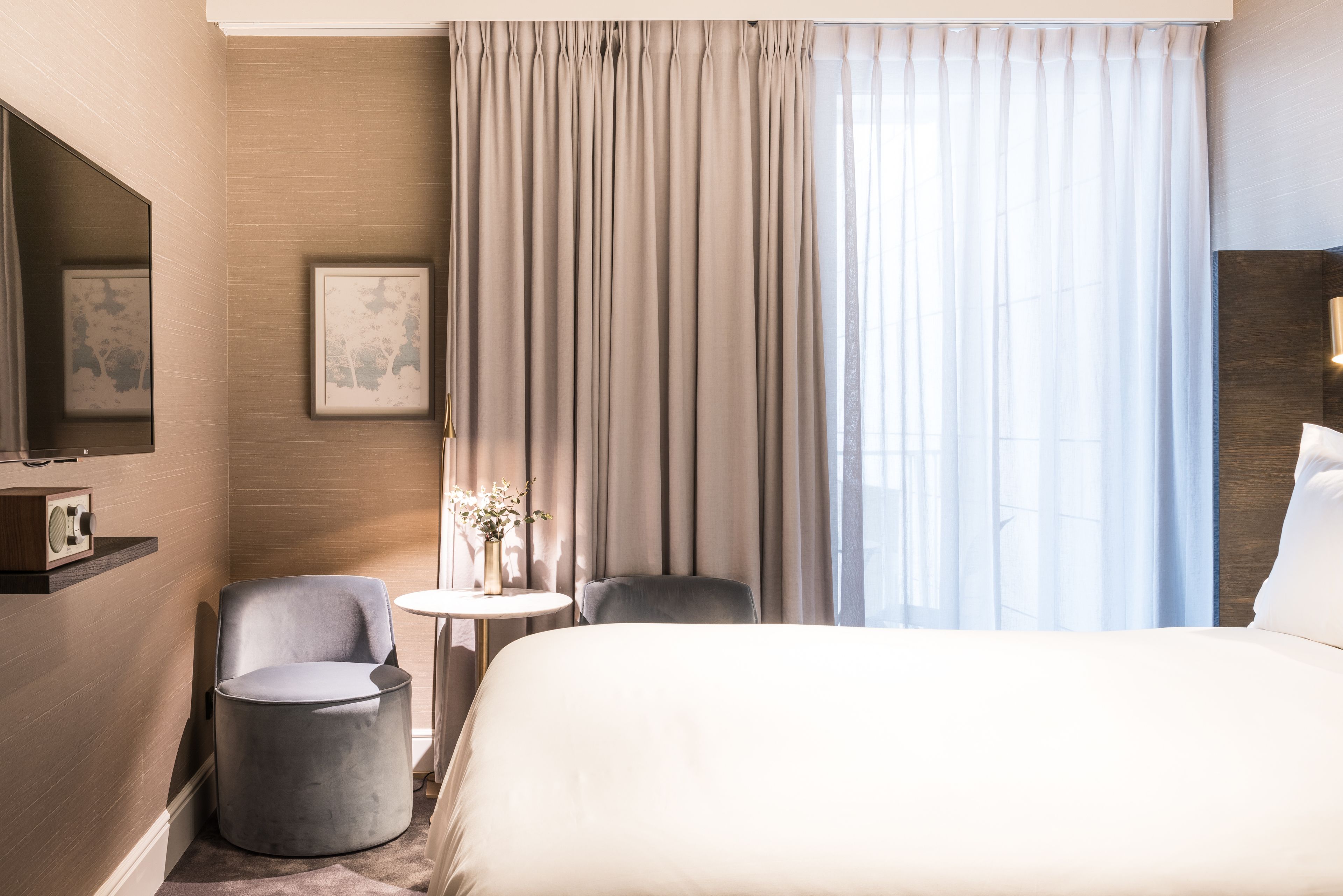 Une chambre d’hôtel avec un grand lit, un fauteuil rond, une petite table avec un vase et une grande fenêtre avec des rideaux.