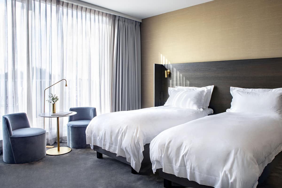 Habitación de hotel con dos camas, una mesita redonda con una lámpara y vistas a la ciudad a través de cortinas translúcidas.