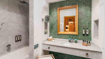 A bathroom with a marble bathtub, a green-tiled vanity, a wooden-framed mirror, and toiletries on the sink.