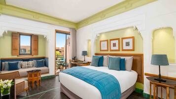 Une chambre d’hôtel avec un grand lit, un canapé, une petite table et une chaise. Il y a une fenêtre avec des volets, un cadre photo accroché au mur et une lampe sur la table de chevet.
