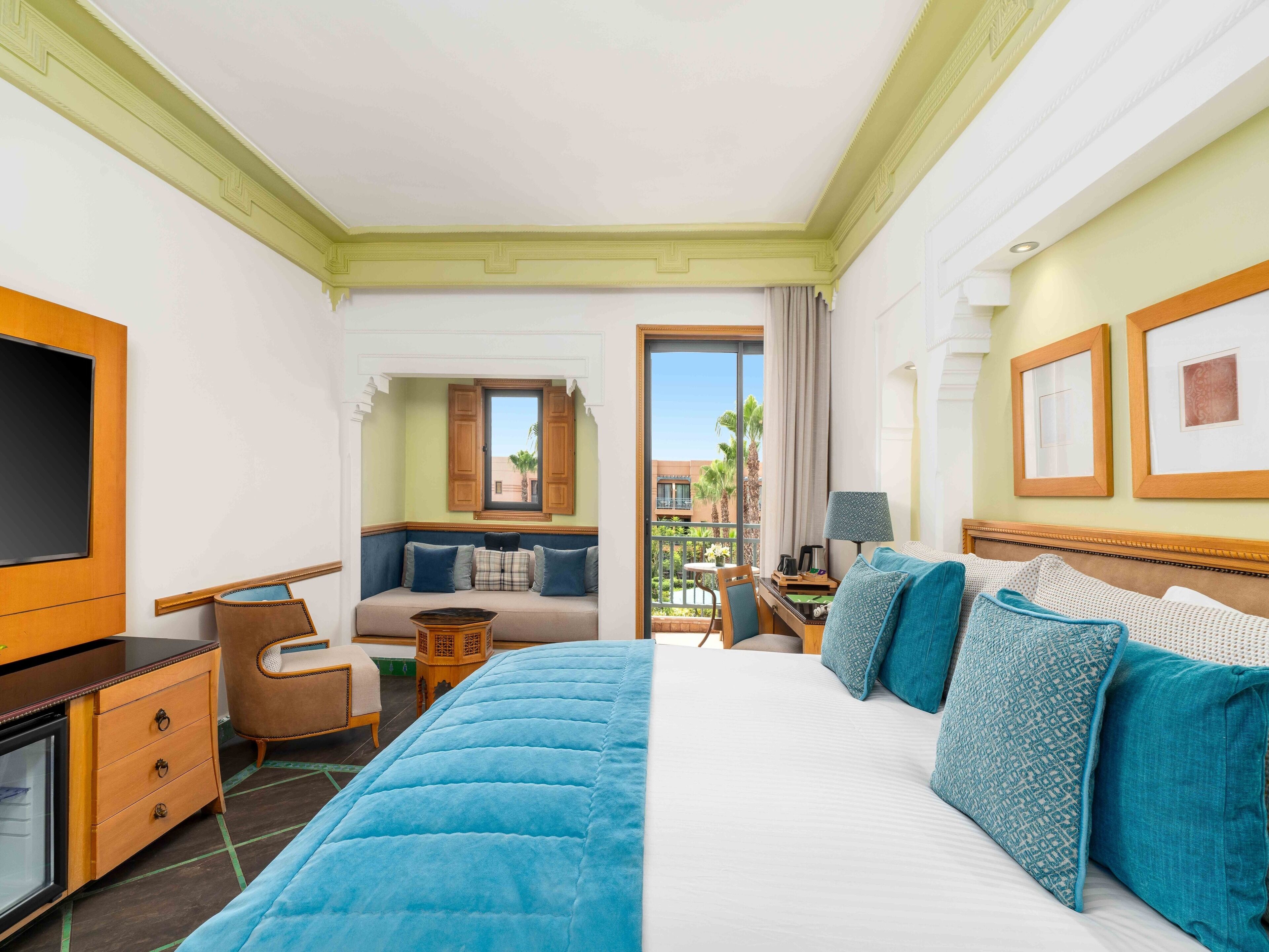 A hotel room with a large bed, a desk, a TV, a balcony with a view of palm trees and buildings.