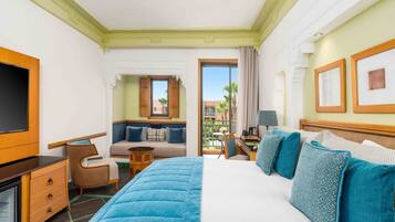 A hotel room with a large bed, a desk, a TV, a balcony with a view of palm trees and buildings.