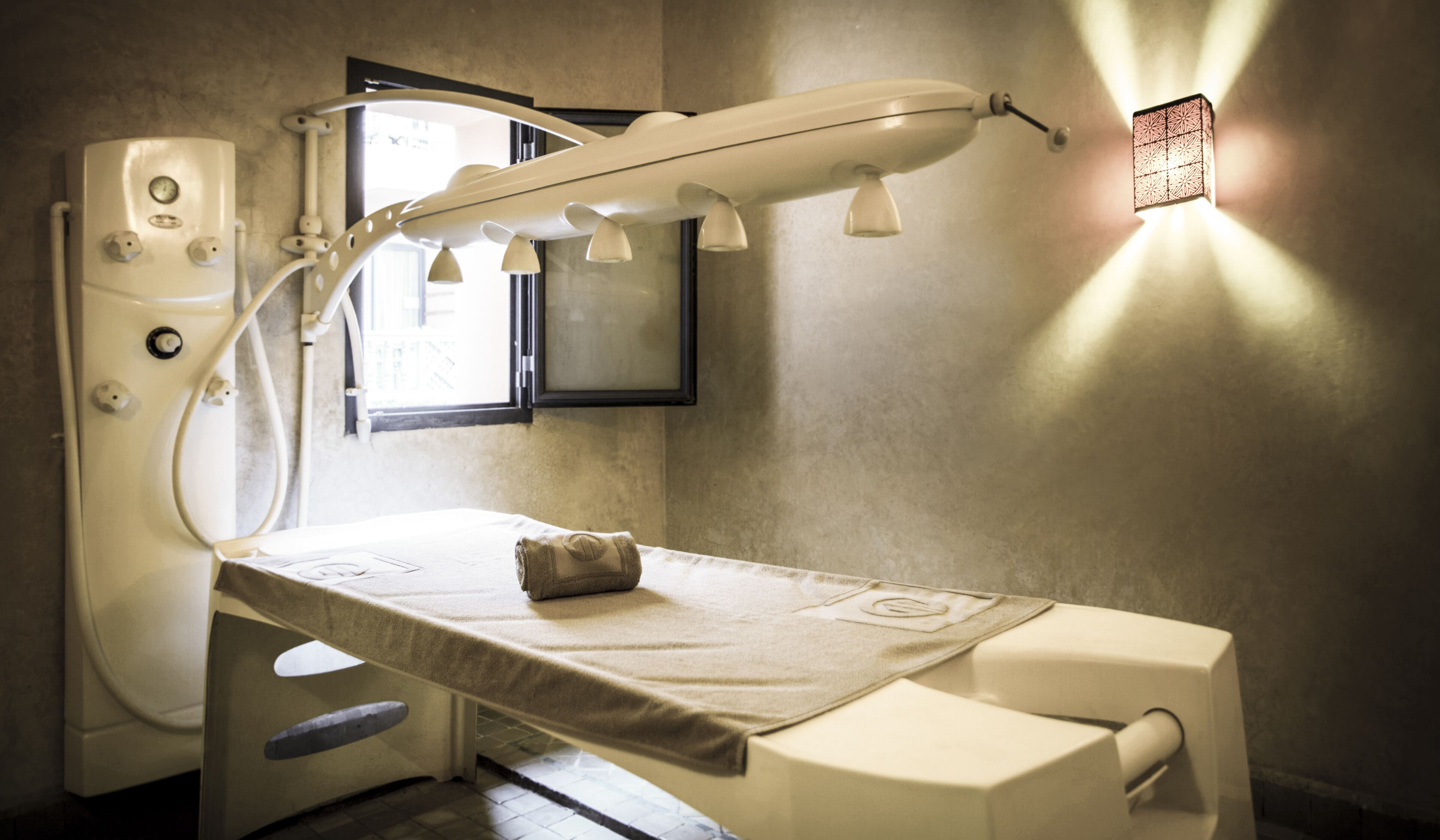 A clinical examination room with a medical examination table, a wall-mounted examination light, a window, and a framed picture on the wall.