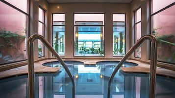 An indoor swimming pool with glass walls and metal railings, surrounded by windows and plants.