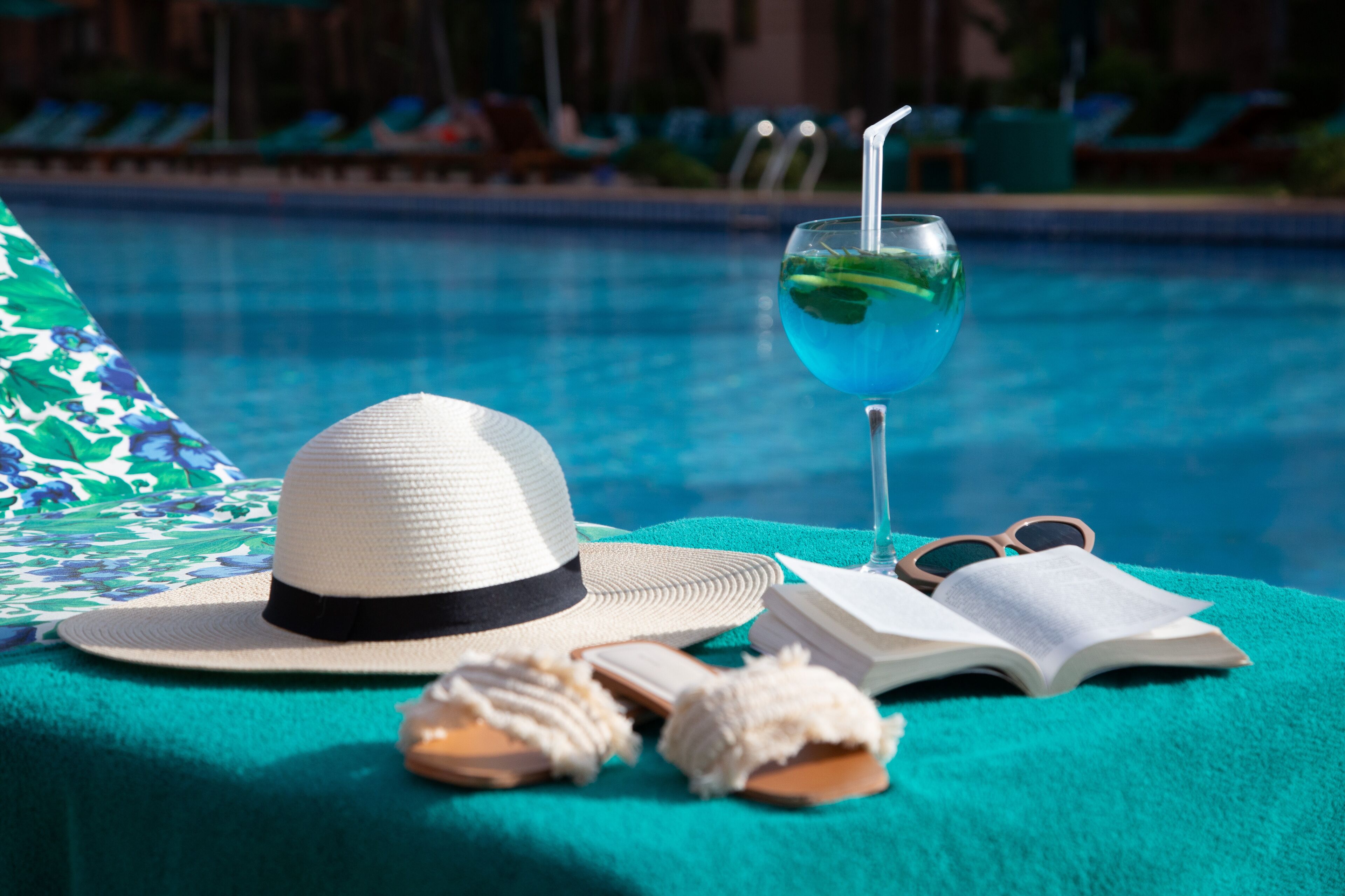 A poolside setting with a hat, sunglasses, a book, and a glass of blue drink on a towel.