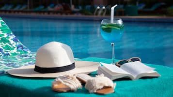 A poolside setting with a hat, sunglasses, a book, and a glass of blue drink on a towel.