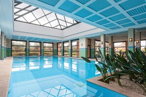 Una amplia piscina interior con techo de tragaluces, rodeada de ventanas y pilares.
