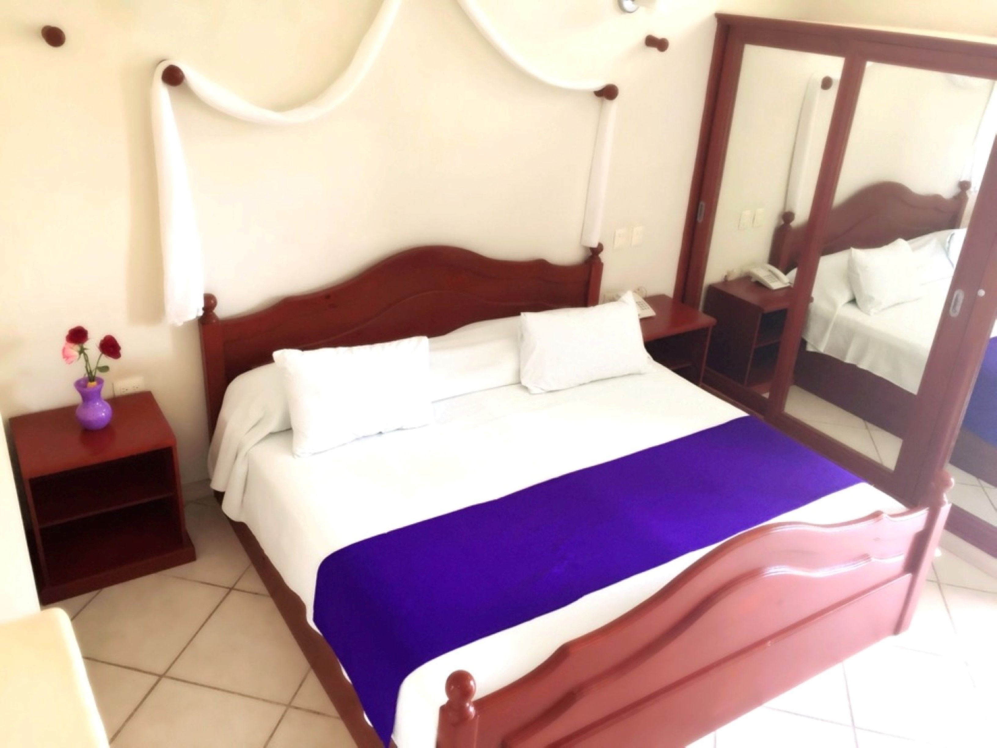 Quarto com cama de madeira, roupa de cama branca, manta roxa, mesinha de cabeceira com um vaso de flores e espelho na parede.