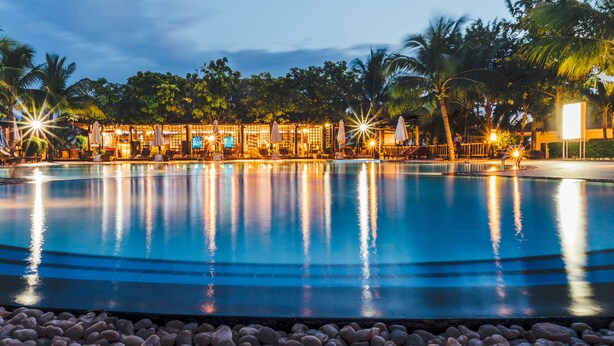 A swimming pool with lounge chairs and umbrellas, surrounded by palm trees and a building with illuminated windows. - Lotus Muine Resort & Spa (Phu Thuy)