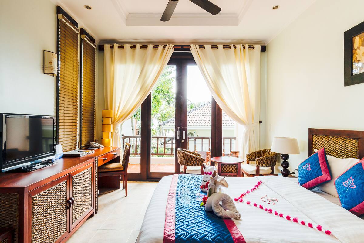 A hotel room with a bed, a television, a desk, and a balcony with chairs.