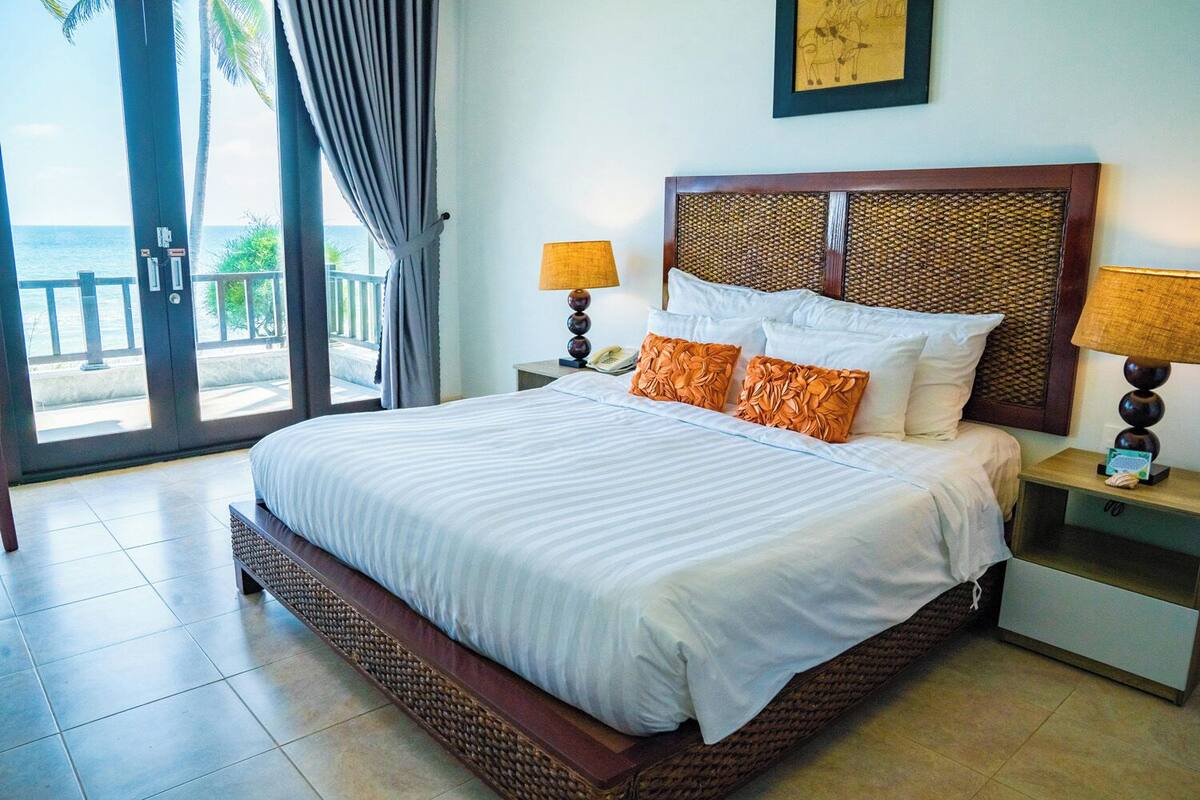 A hotel room with a large bed, two bedside tables with lamps, a view of the ocean through a sliding door, and a framed picture on the wall.