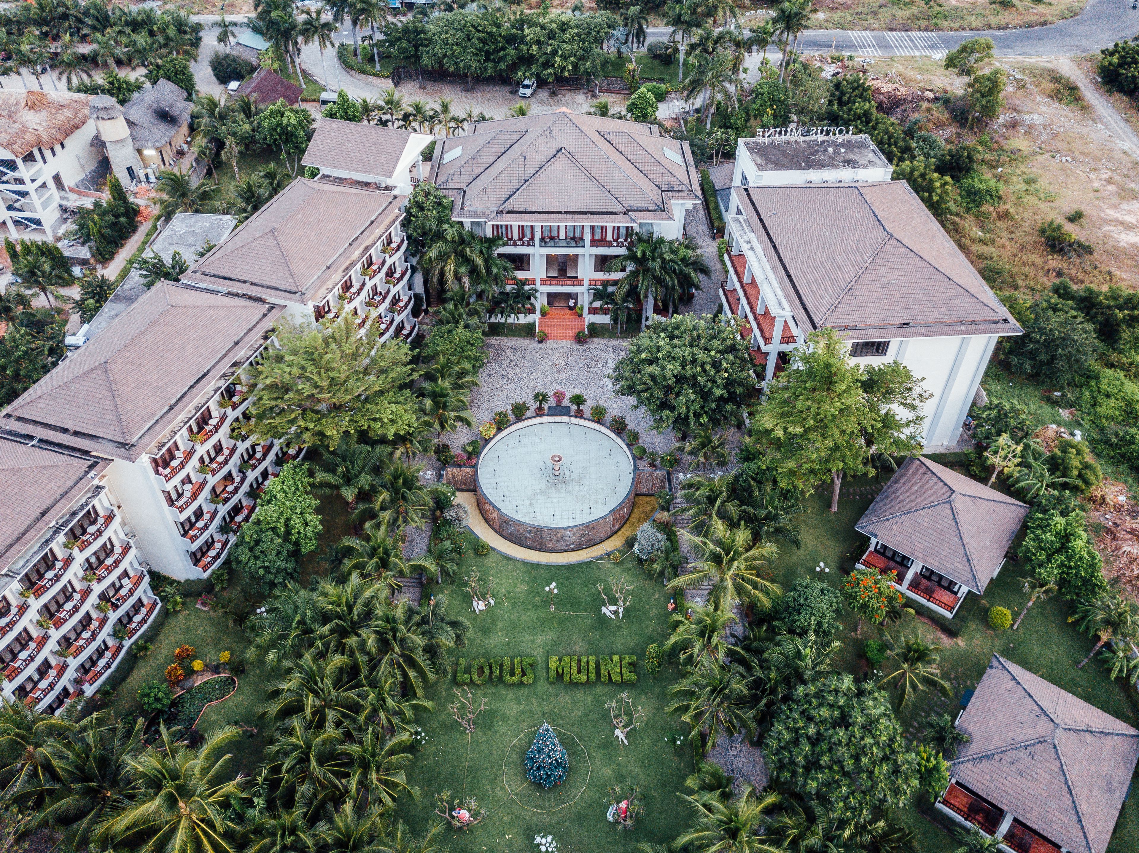 An aerial view of a hotel with multiple buildings, a central courtyard with a circular fountain, and lush greenery.