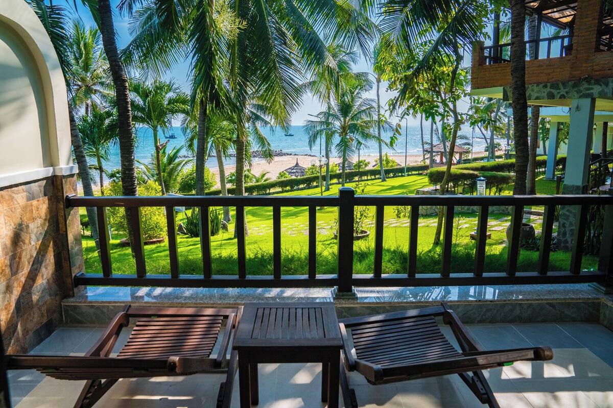 A balcony with wooden chairs and a view of a beach and palm trees.