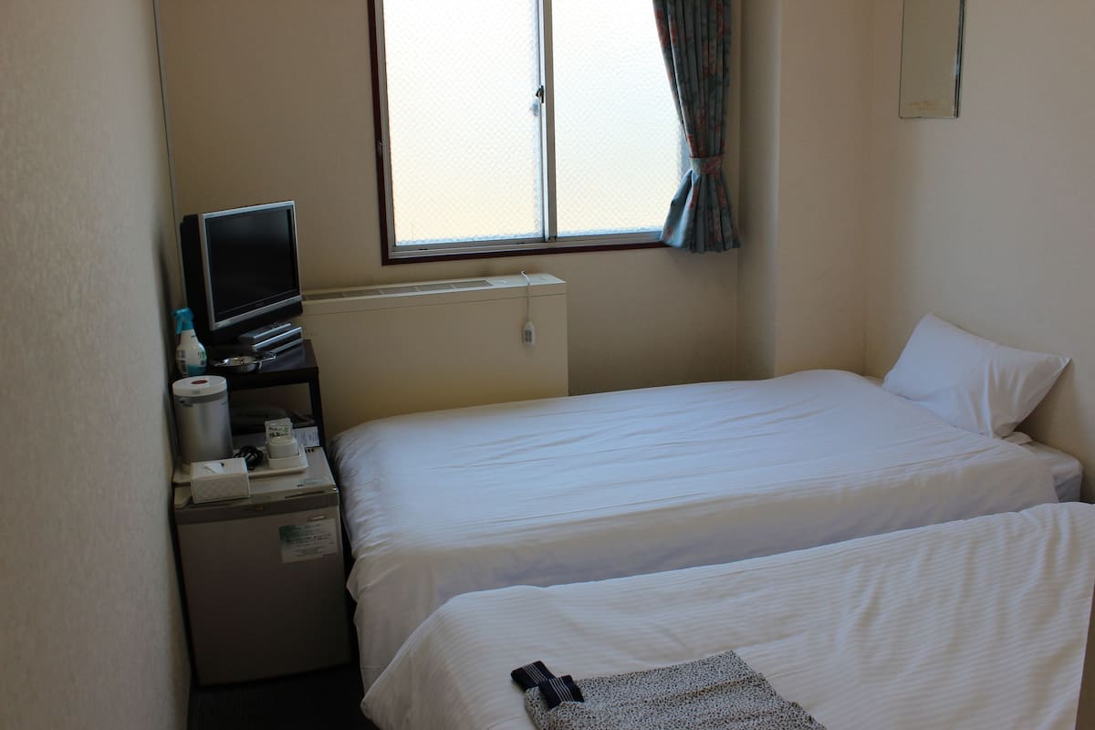 Une chambre d’hôtel avec deux lits, une télévision, un petit bureau avec une lampe et une fenêtre avec des rideaux.