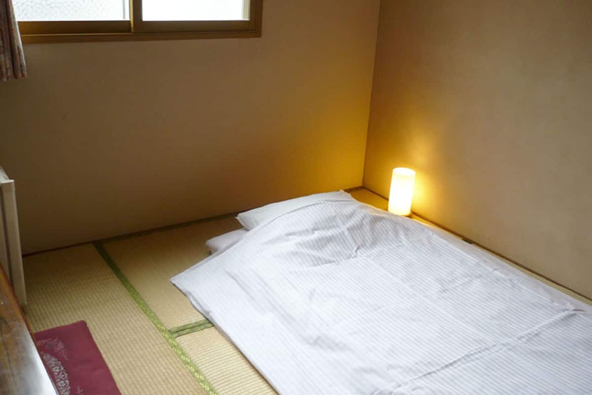 Une petite chambre avec un lit simple, une fenêtre, une lampe de chevet et un futon.
