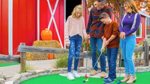 A family playing mini-golf in front of a red barn with hay bales and pumpkins. - Grand Country Waterpark Resort (Branson)