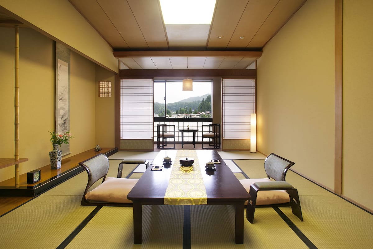 A traditional Japanese room with tatami flooring, a low table with a table runner, and a view of a mountainous landscape through large windows.