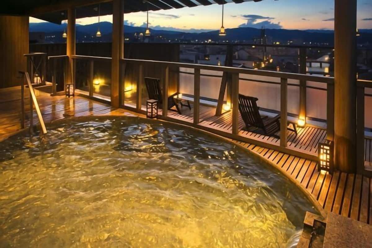 A hot tub on a wooden deck with chairs and tables, overlooking a cityscape at dusk.