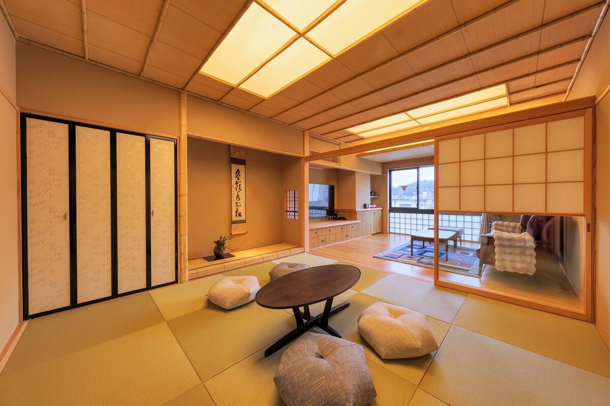Ein traditioneller japanischer Raum mit Tatami-Boden, einem niedrigen Tisch, Kissen, Schiebetüren und Blick durch eine Glaswand.