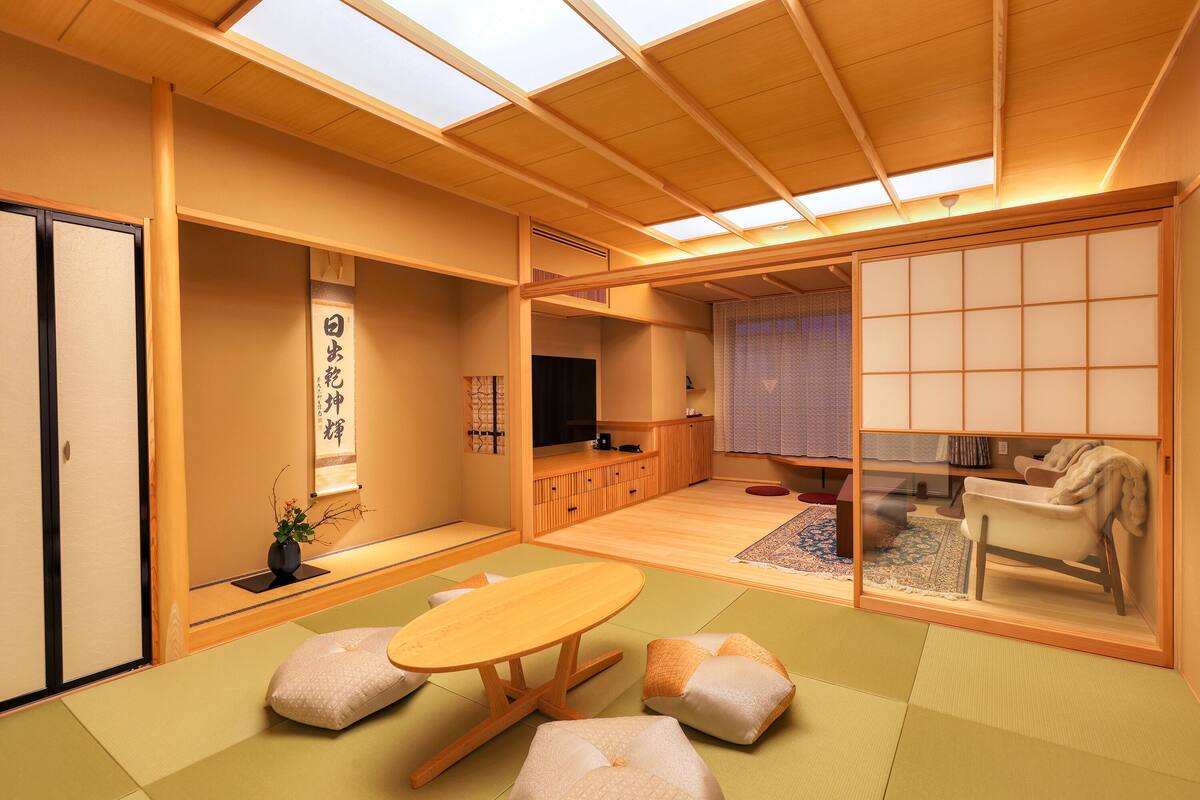 Ein traditioneller japanischer Raum mit Tatami-Bodenbelag, einem niedrigen Tisch, Kissen, Schiebetüren und einem Fernseher.