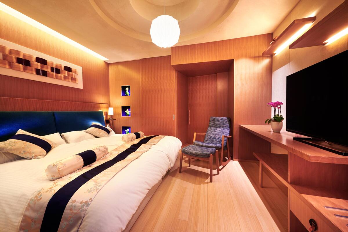 A modern hotel room with a large bed, a seating area, a flat-screen TV, and wooden paneling.