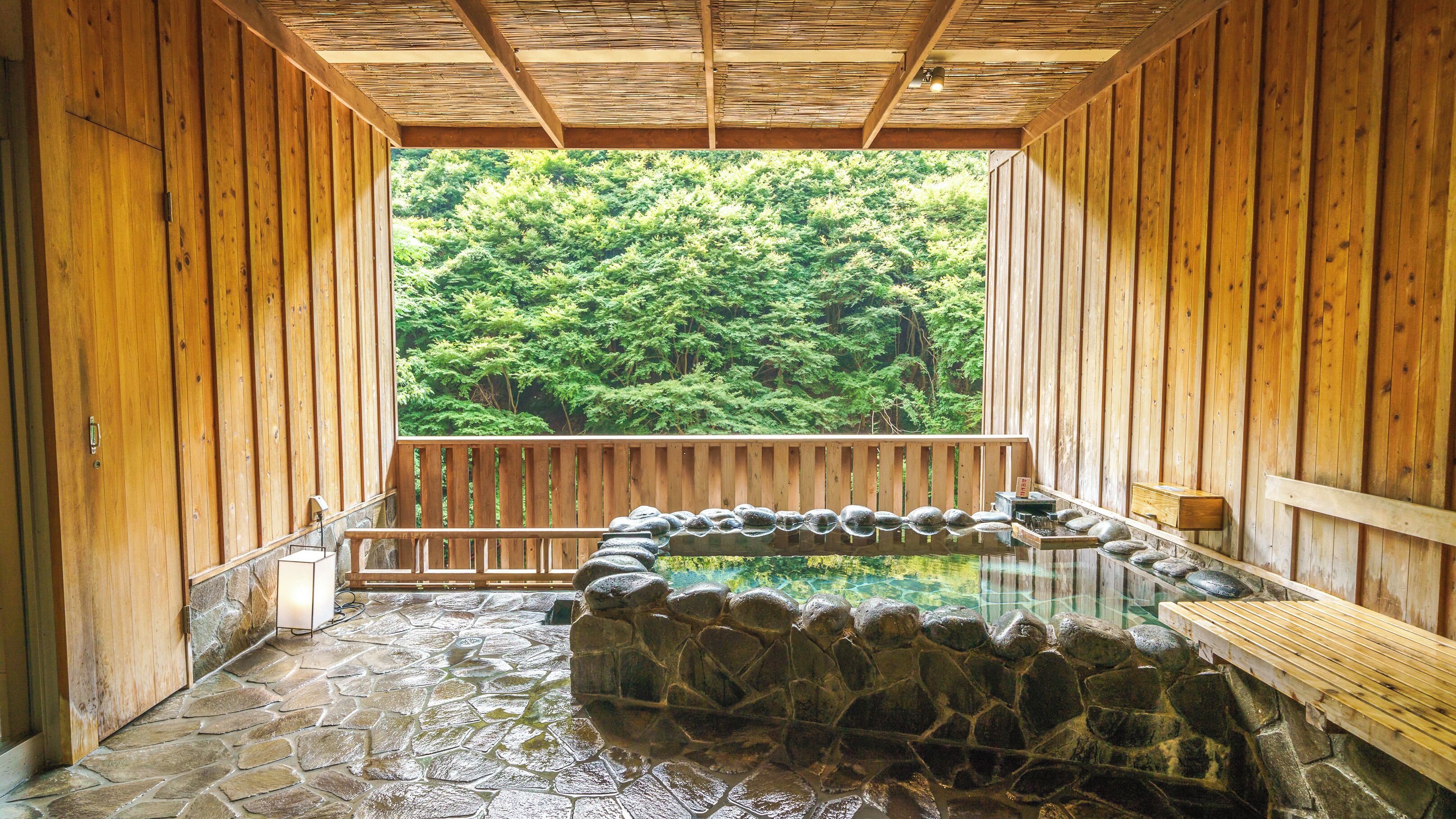 A traditional wooden bathhouse with a stone hot spring, wooden benches, and a view of a forest.