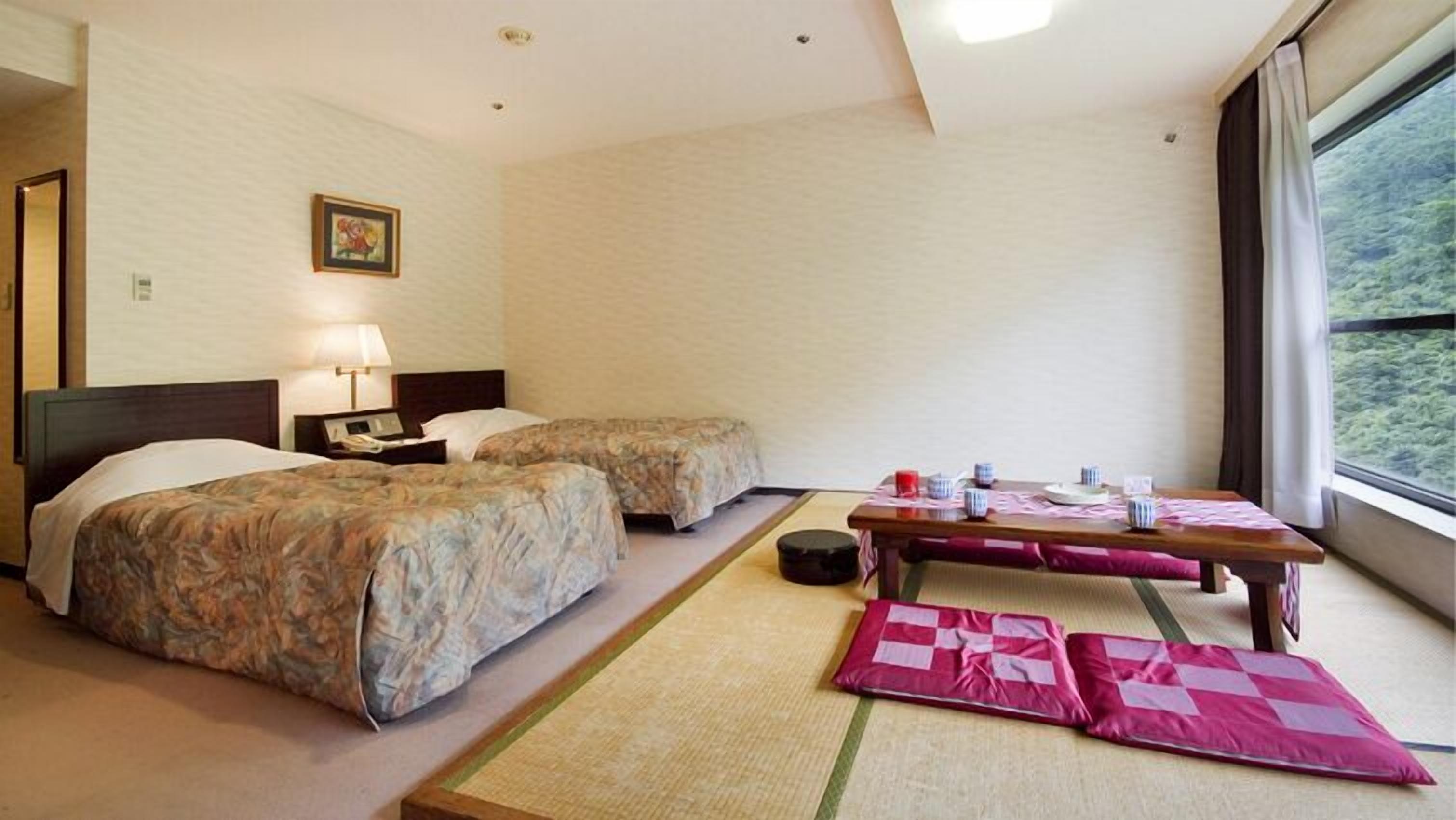 A hotel room with two beds, a bedside table with a lamp, a small table with cups and a plate, and a window with a view of greenery.