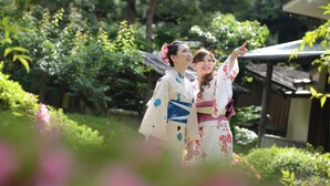 Deux personnes, vêtues de costumes traditionnels, se tiennent dans un jardin, avec une structure en bois en arrière-plan.