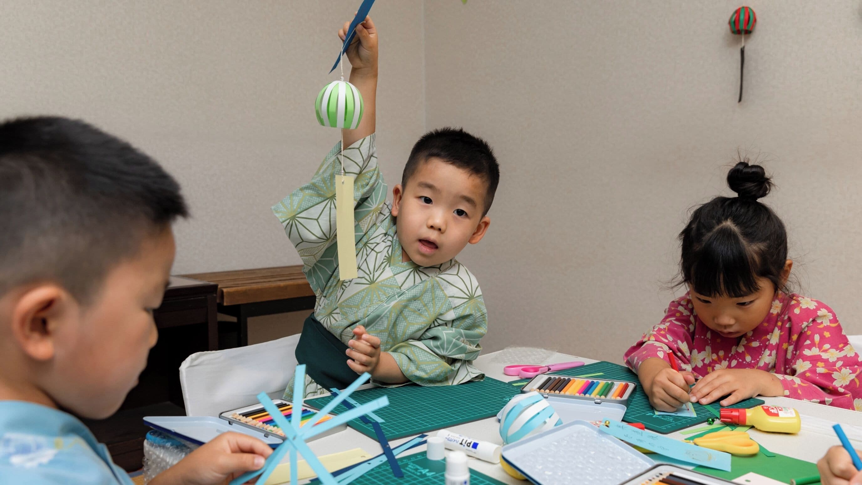 Un bambino che tiene un giocattolo a righe blu e verdi, con un tavolo pieno di materiali artistici e fogli davanti.