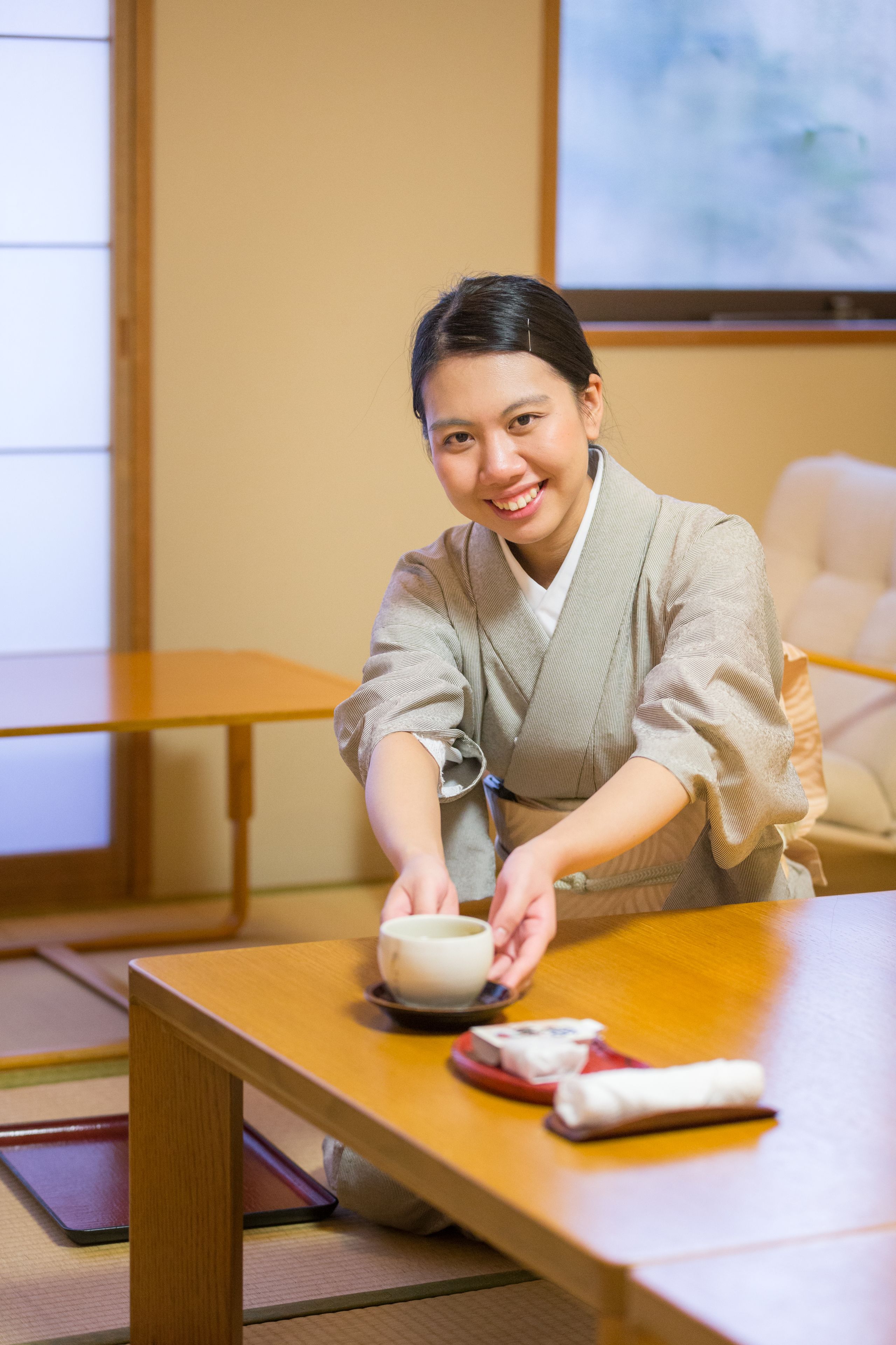 A person in traditional attire sitting at a wooden table with a bowl and chopsticks, in a room with tatami mats and a projector screen in the background.