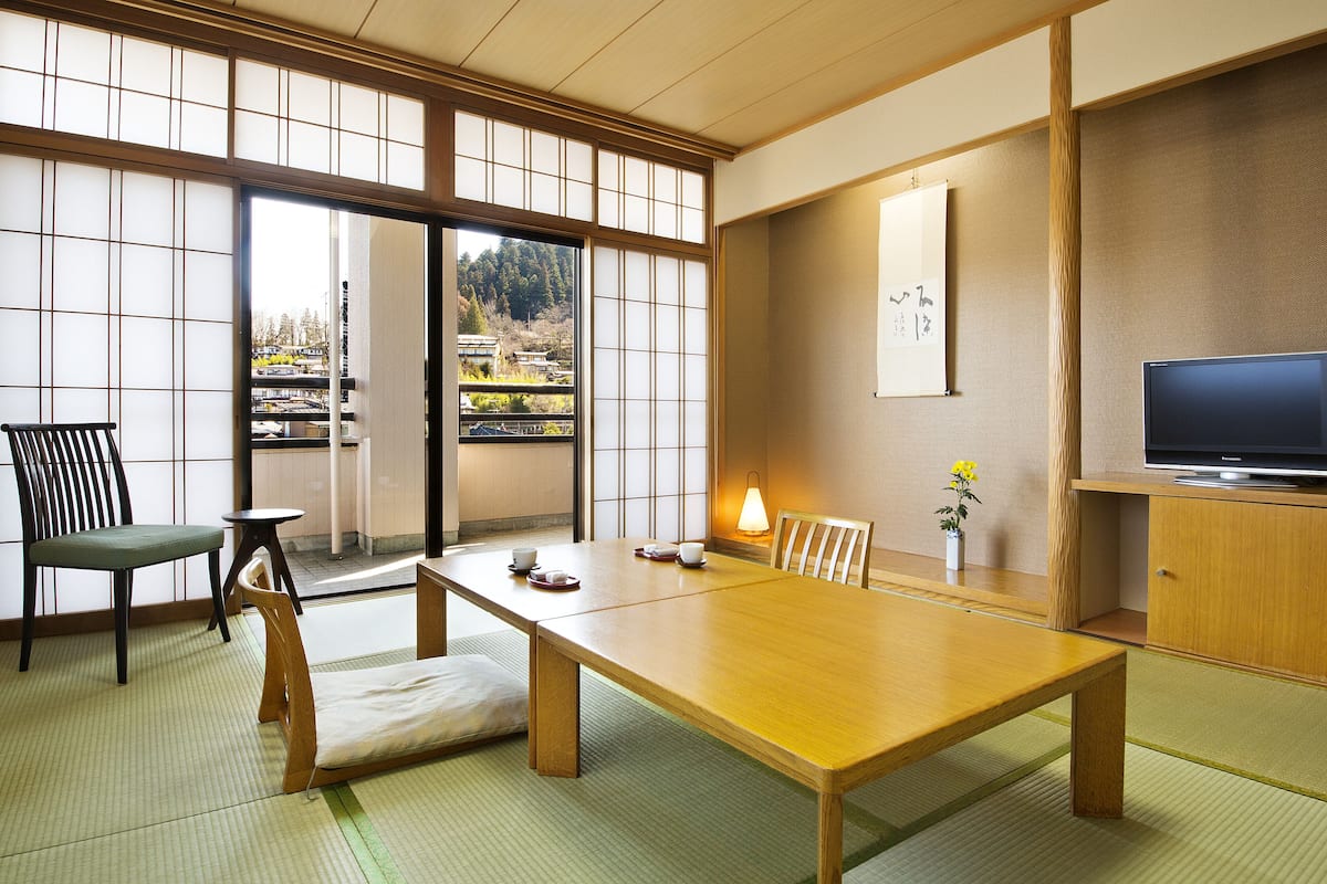 Una habitación japonesa tradicional con suelo de tatami, una mesa baja, sillas, un televisor y vistas a un bosque a través de un puertas correderas.
