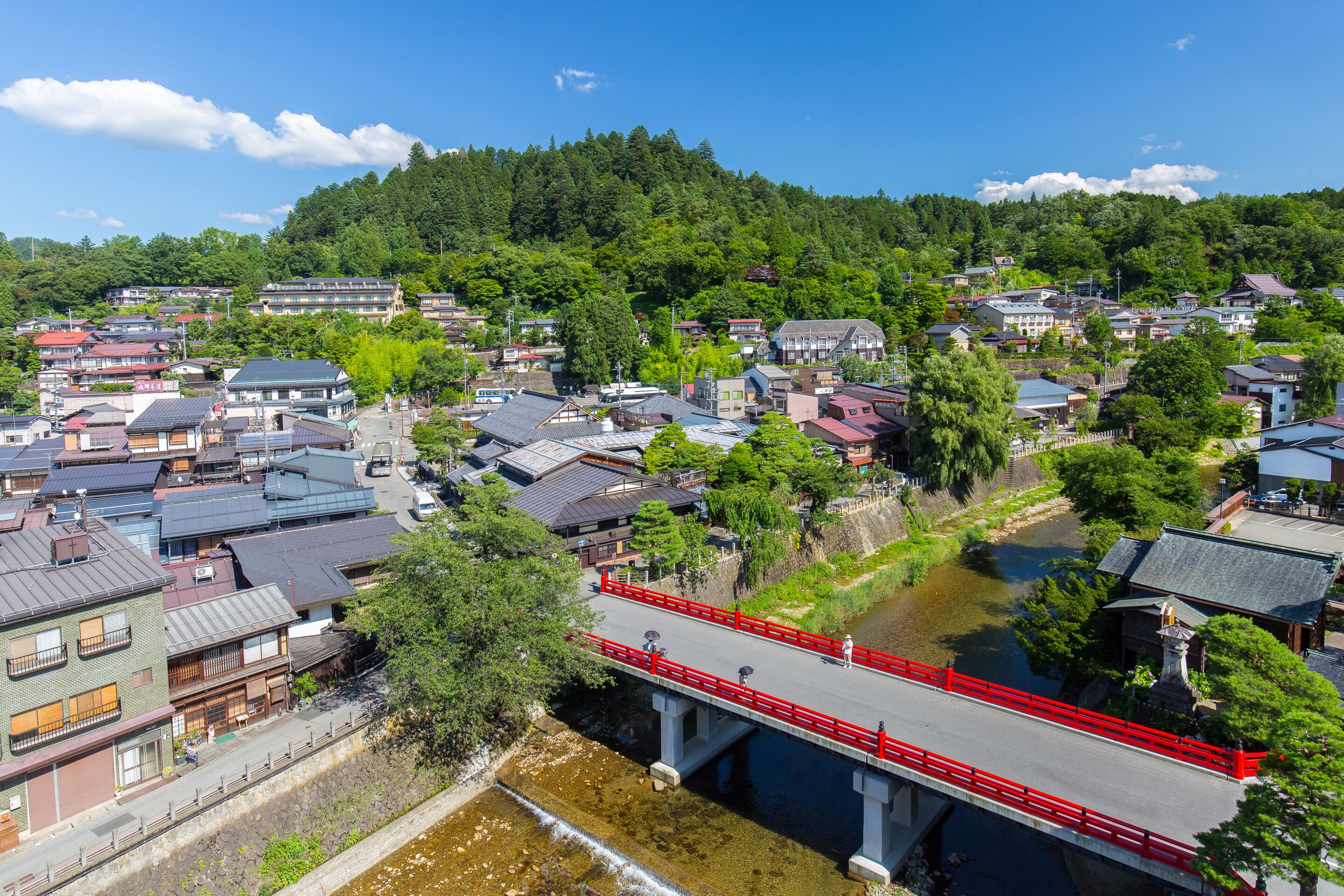 A traditional Japanese town with a river, a red bridge, and buildings with varying roof styles.