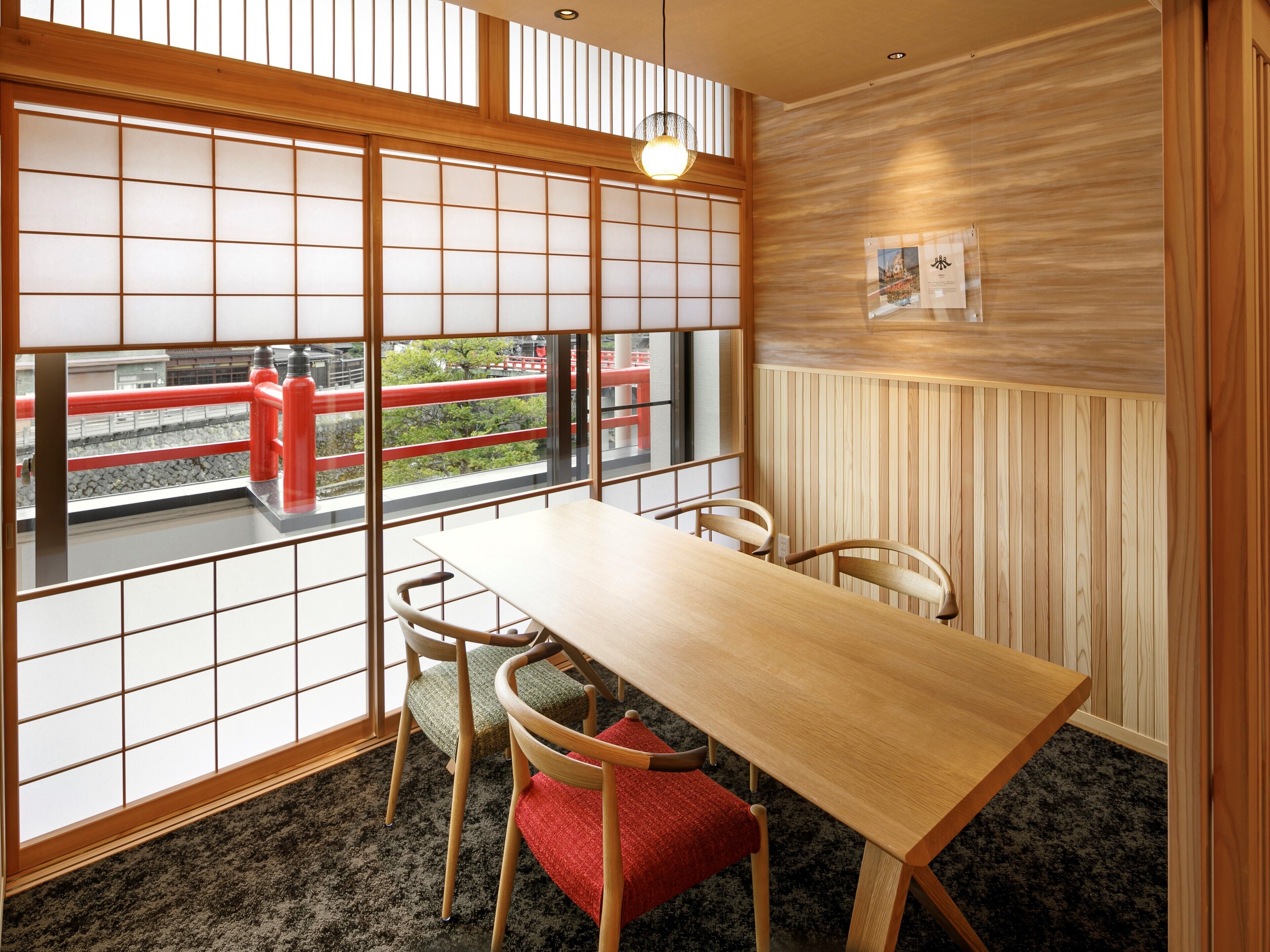 A room with a wooden table and chairs, large windows with a view of a red bridge, and a wooden wall panel.