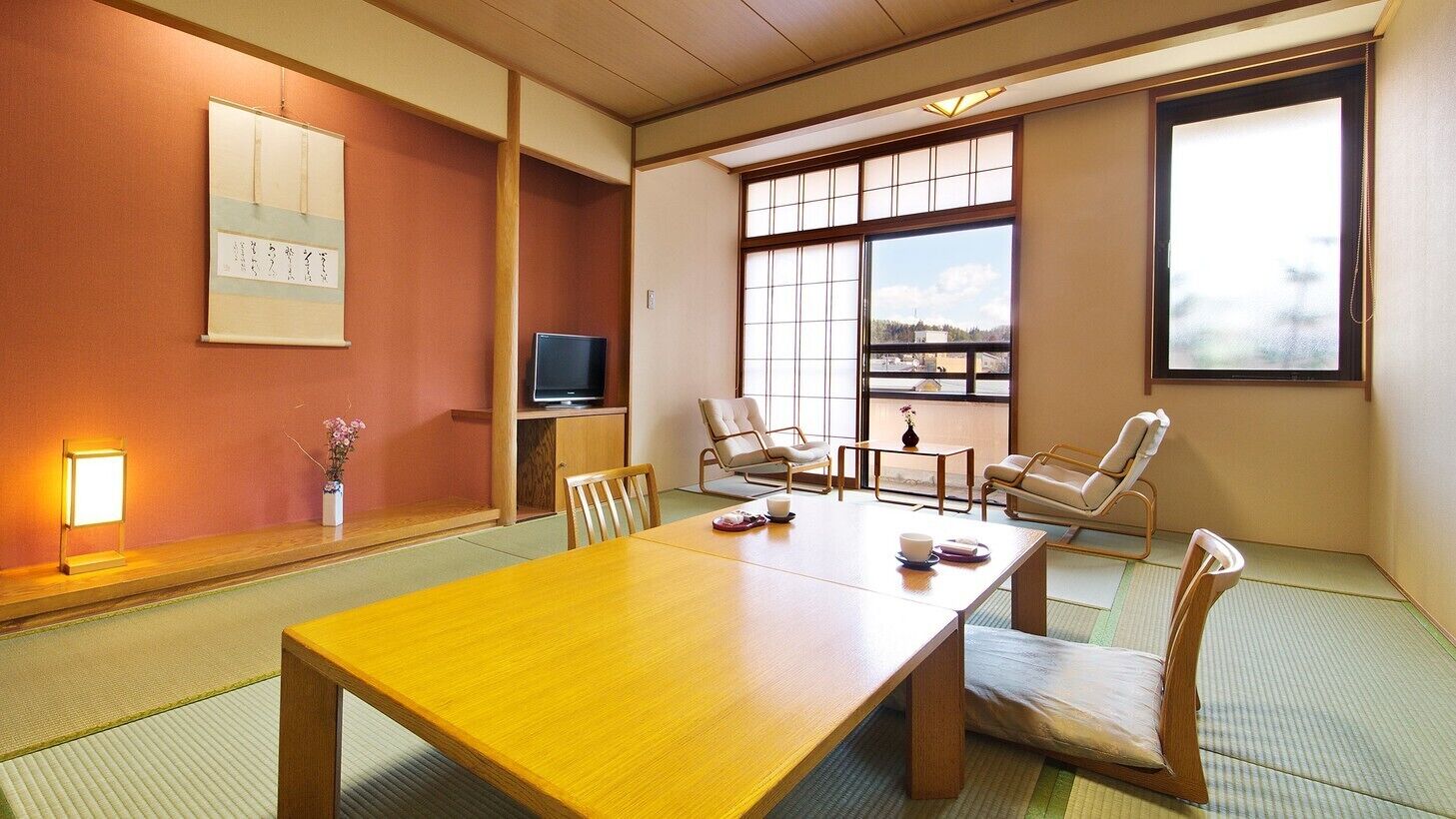 Una habitación japonesa tradicional con suelo de tatami, una mesa de madera, sillas, un televisor y un ventanal con vistas.