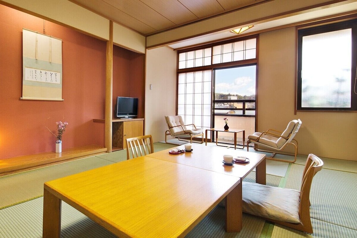Una habitación japonesa tradicional con suelo de tatami, una mesa de madera, sillas, un televisor y un ventanal con vistas.
