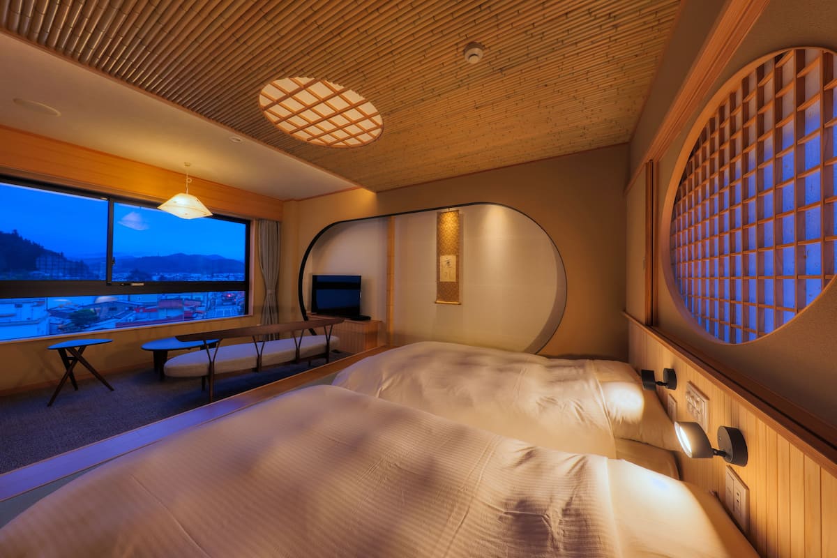 Una habitación de hotel moderna con una cama grande, un televisor y vistas a un paisaje urbano a través de amplios ventanales.