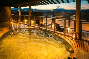 Un bain à remous avec une terrasse en bois et des chaises, offrant une vue sur le paysage urbain au crépuscule.