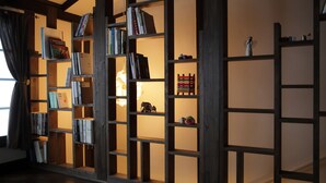 A room with a wooden bookshelf filled with books and decorative items, a small table with a lamp, and a window with curtains. - Gagakuan (Kyoto)