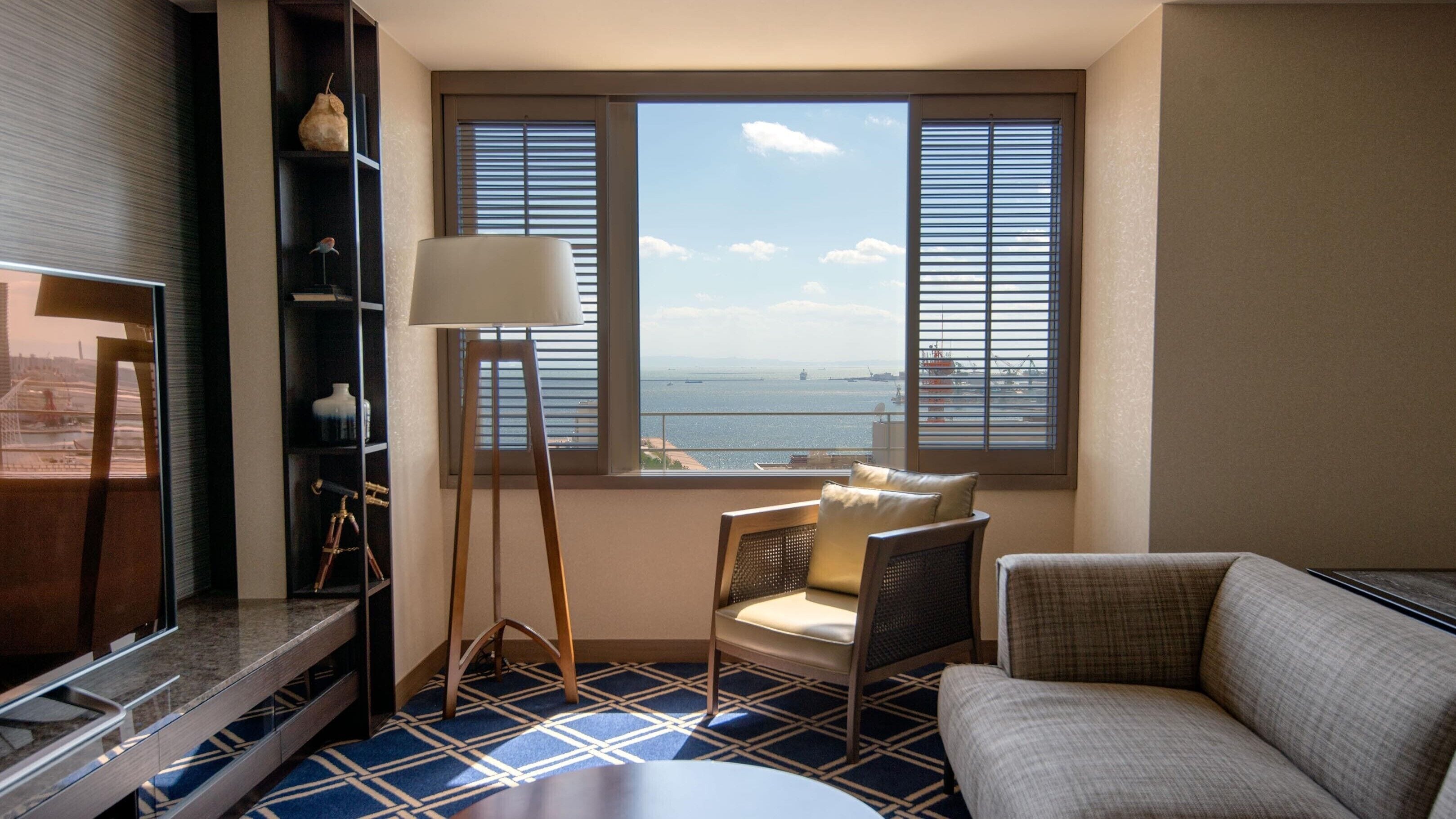 A hotel room with a view of the sea, a sofa, a chair, a lamp, and a shelf with decorative items.