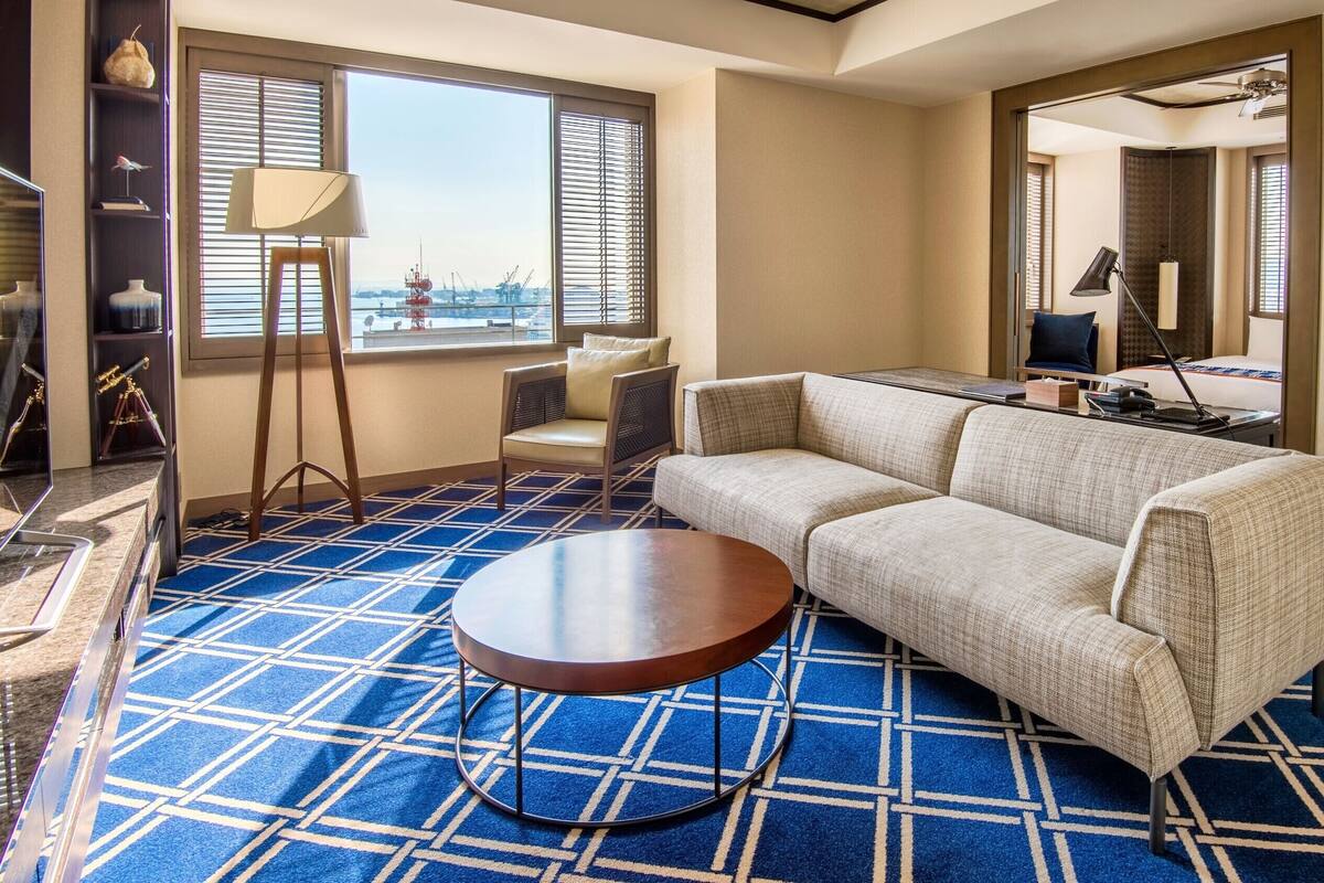 Una sala de estar moderna con alfombra azul estampada, un sofá color beige, una mesa de centro redonda de madera y un ventanal con persianas.