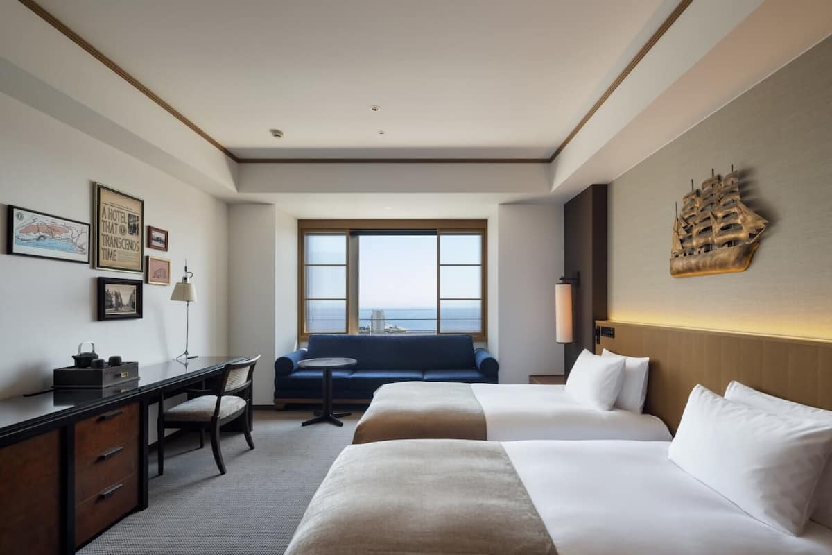 Habitación de hotel con dos camas, un escritorio, una silla, un sofá y un amplio ventanal con vista al mar.