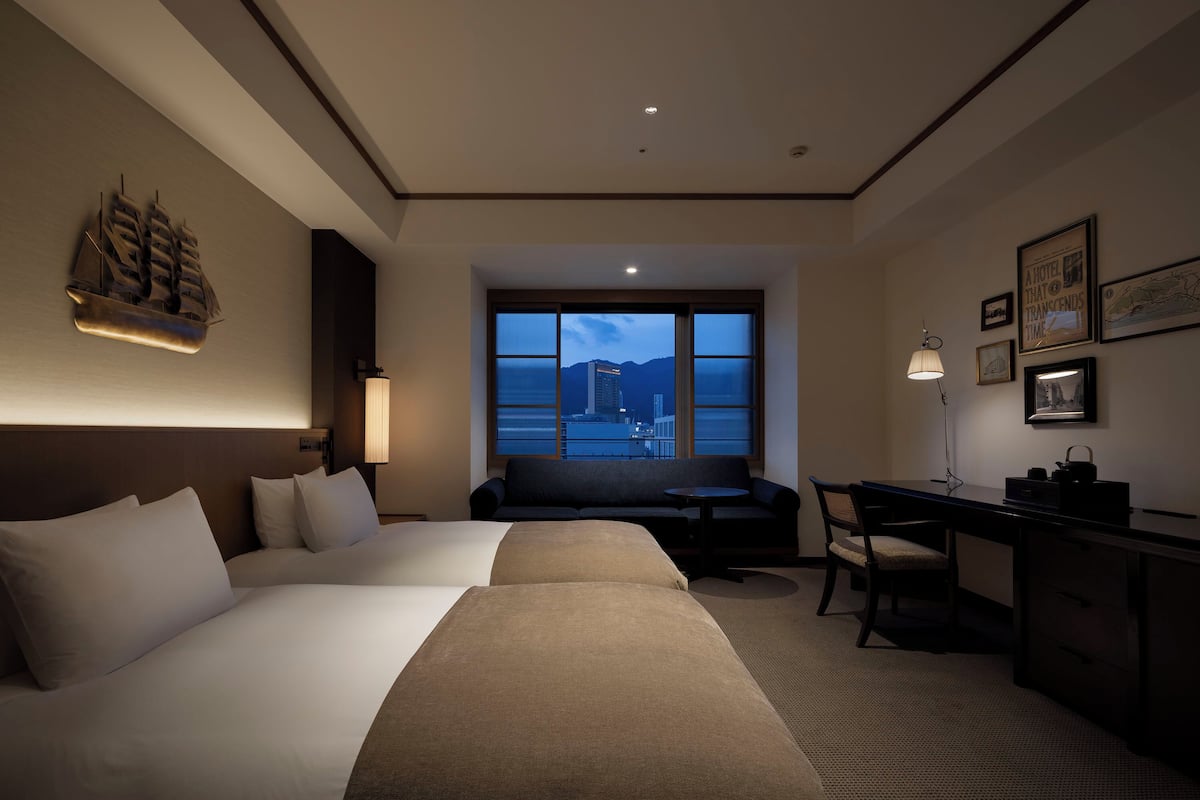 Habitación de hotel con dos camas, un escritorio, una silla, un ventanal con vistas a la ciudad y una decoración náutica en la pared.