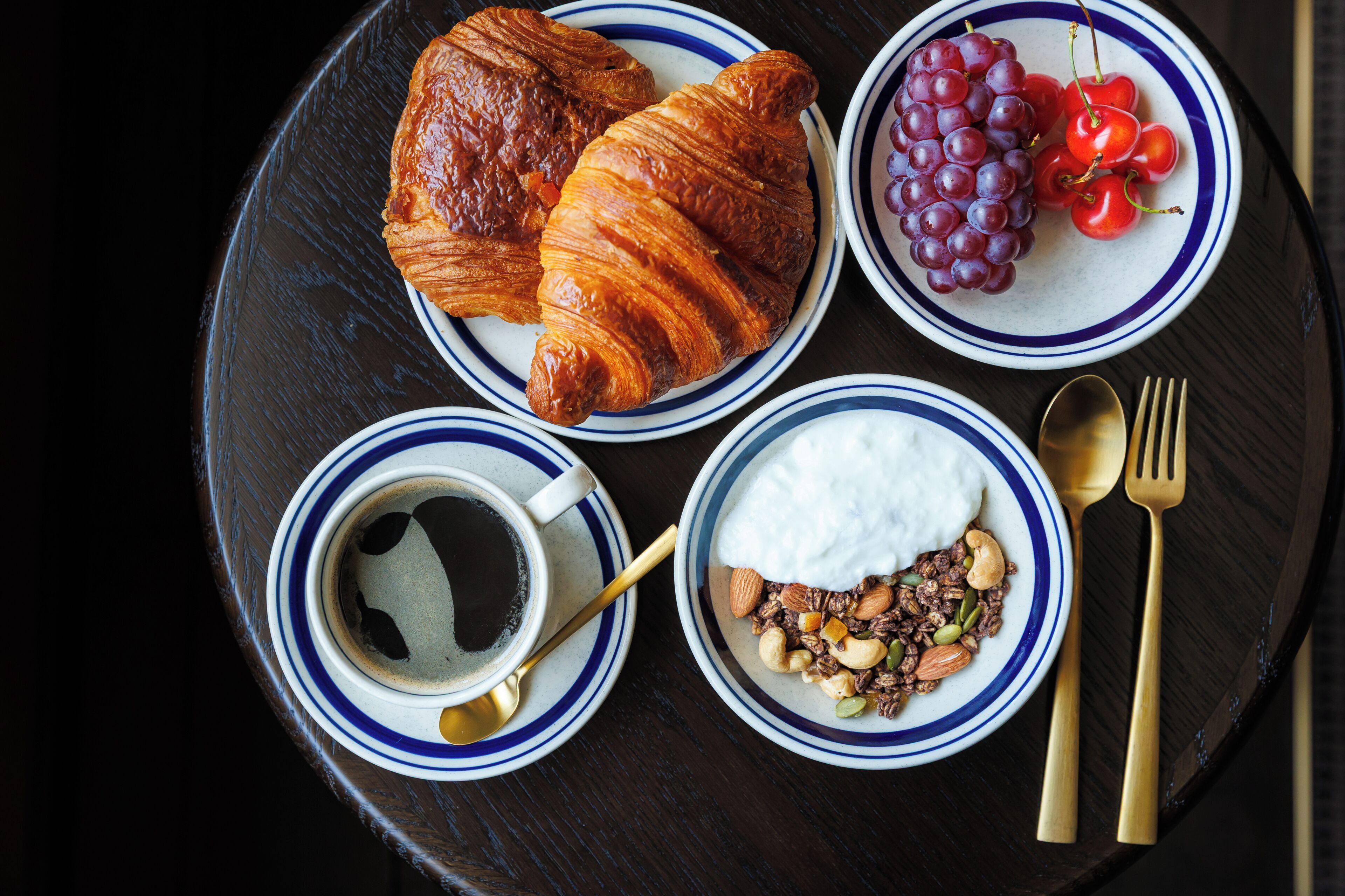 A breakfast tray with croissants, grapes, cherries, yogurt with nuts, and coffee.