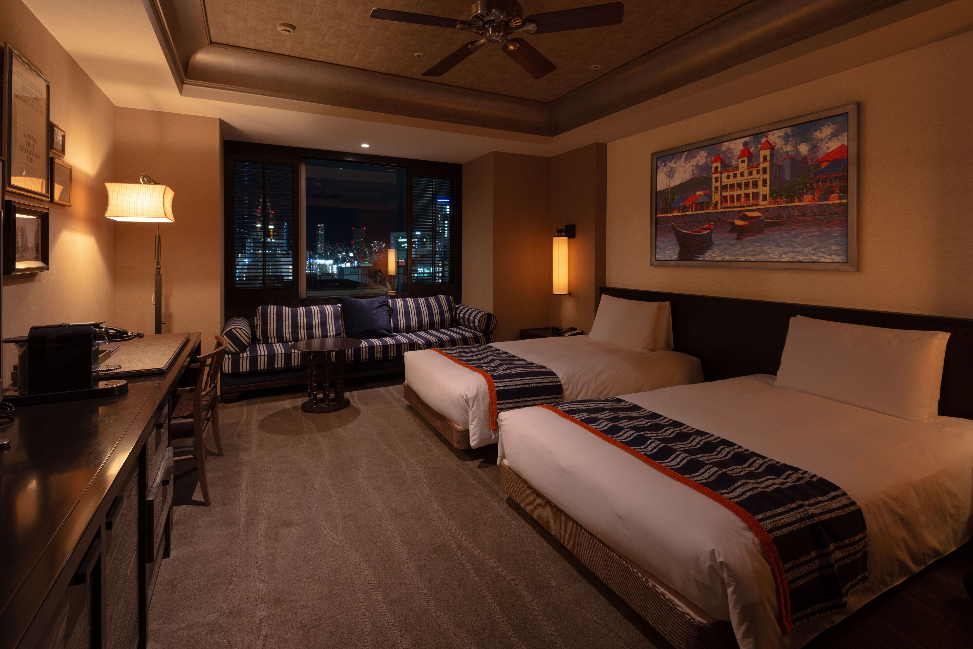 A hotel room with two beds, a desk, a chair, a lamp, a painting, and a view of the city at night.